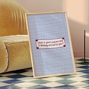 May include: A framed print with a light blue background and a grid pattern. The print features the text "what if your future self is already proud of you?" in a red and white rectangular box. The frame is a light wood color.