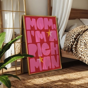 May include: A framed art print with the text "MOM, I'M A RICH MAN" in pink bubble letters on a red background. The print has a gold frame and is leaning against a wall in a bedroom setting.