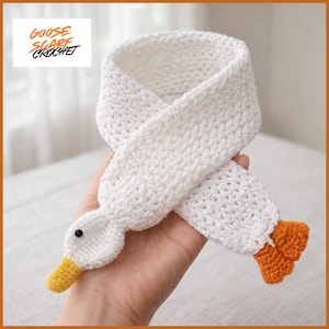 May include: A white crochet scarf shaped like a goose, featuring a yellow beak, black eye, and orange feet. The scarf is handmade and has the text "Goose Scarf Crochet" in the upper left corner.