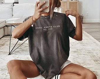 May include: Dark gray t-shirt with the text "COOL AUNTS CLUB EST. 2026" in white. The shirt is a relaxed fit and appears to be made of a soft material. The person is wearing white shorts and white socks with black stripes.