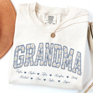 Puede incluir: Una camiseta color crema con la palabra GRANDMA en letras grandes de bloque con rayas azules y un patrón de margaritas blancas. Debajo del texto, están impresos los nombres Taylor, Sophia, Tyler, Meaghan, Jenny, Michael, Sam, Leslie y Jessica.