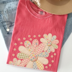 May include: A watermelon-colored t-shirt with a floral applique design. The flowers are made of various patterned fabrics, including gingham and floral prints. The shirt is paired with blue jeans and a tan hat.