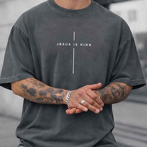 May include: A charcoal gray short-sleeve t-shirt featuring a white vertical cross graphic centered on the chest. The text "JESUS IS KING" is printed in a clean, sans-serif font across the horizontal bar of the cross design on the front.