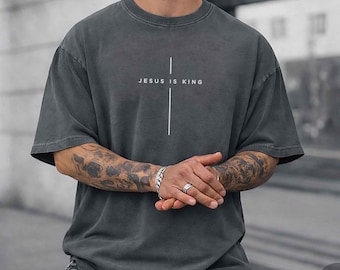 Può includere: Una t-shirt a maniche corte color grigio antracite con una grafica a croce verticale bianca centrata sul petto. Il testo "JESUS IS KING" è stampato con un carattere sans-serif pulito sulla barra orizzontale del disegno della croce.