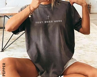 Può includere: Una t-shirt a maniche corte grigio antracite con una piccola icona a croce bianca centrata sopra il testo "TEXT GOES HERE" in un carattere sans-serif pulito. Il capo presenta un girocollo e una silhouette ampia e rilassata.