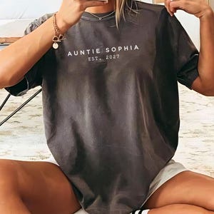 May include: A charcoal grey short-sleeve t-shirt featuring the white text AUNTIE SOPHIA EST. 2027 printed in a clean, sans-serif font across the chest. The garment has a crew neckline and a relaxed, loose-fitting silhouette with short sleeves.