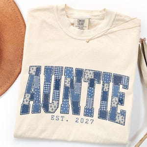 May include: An ivory t-shirt featuring the word AUNTIE in large block letters filled with a blue and white patchwork pattern. Below the word, the text EST. 2027 is printed in blue. The shirt has a Comfort Colors brand label at the inner neckline.