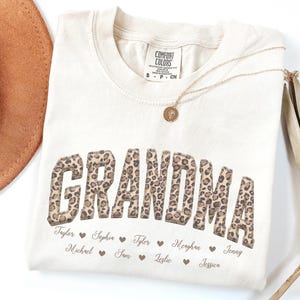 Może przedstawiać: Kremowy t-shirt z krótkim rękawem z napisem GRANDMA w brązowo-czarny wzór w panterkę. Pod tekstem wydrukowano kilka imion kursywą, oddzielonych małymi brązowymi serduszkami. Koszulka posiada metkę z napisem Comfort Colors.
