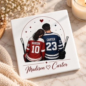 May include: A square white ceramic tile featuring a cartoon illustration of two people in hockey jerseys, one red with the name Madison and number 10, the other blue with the name Carter and number 23, holding hockey sticks with the text Madison Carter.