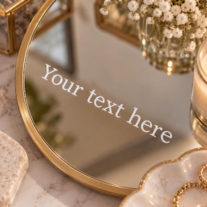 May include: A round mirror with a thin gold-colored metal frame. The surface of the mirror features the white text "Your text here" printed in a clean, sans-serif font. The mirror reflects light and surrounding objects in a bright setting.