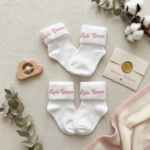 May include: Two pairs of white ankle socks with folded cuffs, each featuring the name Ada Emma embroidered in pink script. The socks are displayed alongside a wooden cloud-shaped rattle and a small envelope sealed with a gold wax heart stamp.
