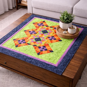 以下が含まれることがあります： 幾何学的なスターバーストデザインのカラフルなキルト。キルトは明るい緑色の中心部を持ち、紫とネイビーブルーのボーダーで縁取られています。オレンジ、紫、黒の正方形が星のパターンを形成しています。キルトは木製のコーヒーテーブルの上にあります。