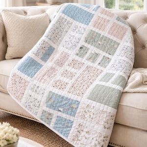 May include: A patchwork quilt with a floral pattern in pastel colors, including blue, pink, and green. The quilt is draped over a beige sofa, creating a cozy and inviting aesthetic. The quilt's design features a variety of floral prints.