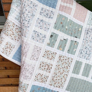 May include: A quilted blanket with a patchwork design. The quilt features rectangular blocks in various floral patterns and solid colors, including blue, pink, and brown, separated by white strips. The text "Designed by PatternNetStudio" is visible.