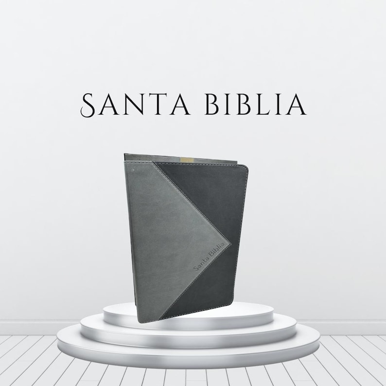 K&ouml;nnte beinhalten: Eine grau-schwarze Bibel mit dem Text "SANTA BIBLIA" dar&uuml;ber. Die Bibel steht auf einem wei&szlig;en, mehrstufigen Pr&auml;sentationsst&auml;nder. Das Cover hat ein geometrisches Design.