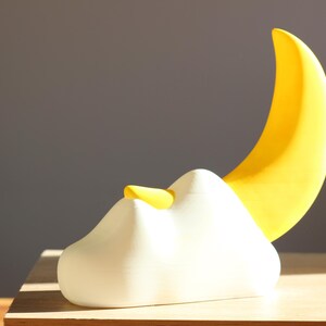 Könnte beinhalten: Eine skurrile Skulptur mit einer weißen wolkenartigen Form mit einem kleinen gelben Akzent und einer großen, gebogenen gelben Halbmondform. Das Kunstwerk befindet sich vor einem neutralen grauen Hintergrund.