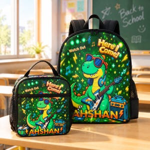 Peut inclure: Un sac à dos et un sac à déjeuner assorti verts ornés d'un dinosaure de dessin animé portant des lunettes de soleil et un casque tout en jouant de la guitare électrique. Le motif inclut le texte "Watch Out Here I Come!" et "AHSHAN".