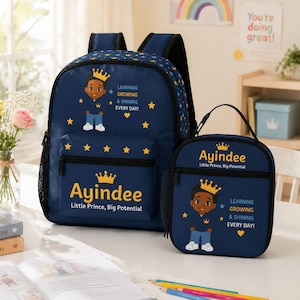 May include: A dark blue backpack and matching lunch bag set featuring a cartoon illustration of a young boy wearing a gold crown. The text reads Ayindee, Little Prince, Big Potential, and Learning Growing & Shining Every Day with yellow stars.