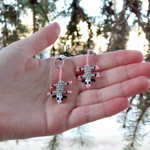 May include: A pair of handmade beaded opossum earrings. Each earring features a gray body, pink feet, and black and white details. The earrings are suspended from a pink beaded chain and a silver-tone hoop.