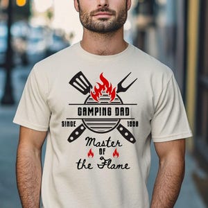 May include: Beige t-shirt with a black graphic that reads "Camping Dad Master of the Flame" with a grill, spatula, and fork design. The text "Personalized with Year" is at the top left.