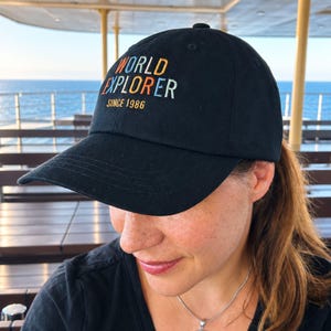 Könnte beinhalten: Eine schwarze Baseballkappe mit dem gestickten Text WORLD EXPLORER in Orange, Hellblau und Weiß sowie dem darunter in Orange gestickten Text SINCE 1986. Die Kappe hat einen gebogenen Schirm und eine strukturierte Krone mit einem Knopf.