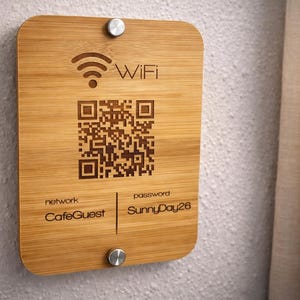 May include: A rectangular bamboo WiFi sign with rounded corners. The sign features a WiFi symbol, a QR code, and the network name "CafeGuest" and password "SunnyDay26". The sign is mounted with two silver screws.