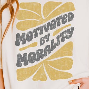 May include: An ivory short-sleeved t-shirt featuring a graphic design with the text MOTIVATED BY MORALITY in dark grey letters. The text is layered over a mustard yellow abstract shape with a distressed, weathered texture on the print.
