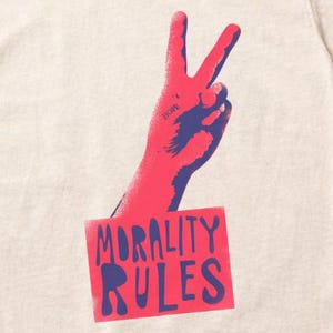 May include: Cream-coloured t-shirt featuring a red and blue graphic of a hand making a peace sign. The text "MORALITY RULES" is printed in red on a rectangular blue background. The shirt is layered over a light blue denim jacket.