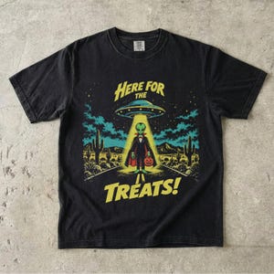 May include: A black short-sleeved t-shirt featuring a graphic of an alien dressed as a vampire standing under a UFO beam in a desert landscape. The text "HERE FOR THE TREATS!" is printed in yellow above and below the central illustration.