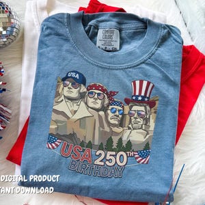 Puede incluir: Una camiseta azul de manga corta con un gráfico de los cuatro presidentes del Monte Rushmore con gafas de sol con patrones de la bandera estadounidense. El texto dice USA 250TH BIRTHDAY. La camiseta tiene una etiqueta Comfort Colors en el cuello.