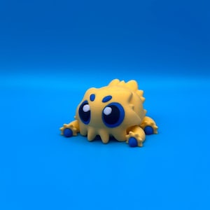 May include: A yellow, 3D-printed creature with large blue eyes and small blue accents. The creature has a spiky back and four small legs. It is set against a solid blue background.