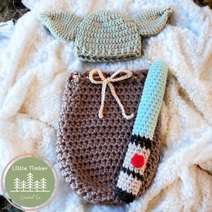 Puede incluir: Un conjunto de ropa de bebé de ganchillo, que incluye un gorro verde claro con orejas grandes, un saco marrón con un cordón crema y un sable de luz azul y blanco. El conjunto está diseñado para parecerse a un personaje popular.