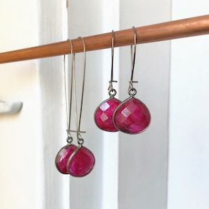 May include: A pair of dangle earrings with pink gemstone drops. The earrings have silver hooks and are hanging on a wooden dowel.