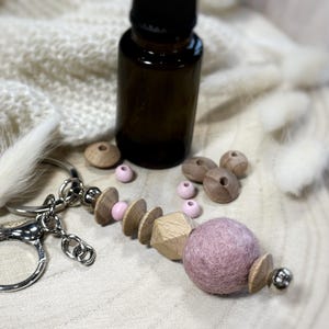 Puede incluir: Un llavero plateado con un diseño decorativo de cuentas de madera y rosas. El llavero presenta una bola de fieltro malva grande y redonda y varias cuentas de madera. Una pequeña botella de vidrio marrón y otras cuentas de madera también están en la imagen.