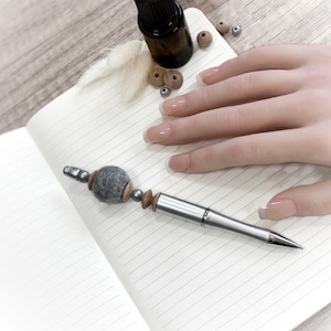 Op de afbeelding: Een zilveren pen met een grijze vilten bal en houten kralen ligt op een gelijnd notitieboek. Een kleine bruine fles en houten kralen liggen ook op het notitieboek. Een hand met verzorgde nagels rust op het notitieboek.