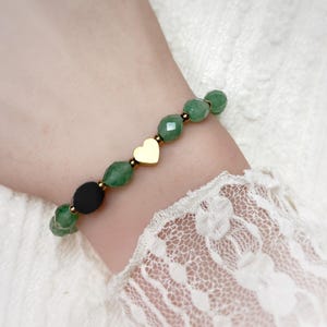 Könnte beinhalten: Ein Perlenarmband mit facettierten grünen Edelsteinperlen, einer einzelnen glatten schwarzen runden Perle und einem zentralen goldfarbenen herzförmigen Anhänger. Die Perlen sind durch kleine metallische goldfarbene Zwischenstücke auf einer Schnur getrennt.