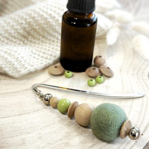 May include: A silver metal bookmark featuring a decorative charm composed of a large sage green felt ball, natural wooden beads in varying shapes, and small metallic silver spheres. The bookmark is positioned alongside a dark amber glass bottle.