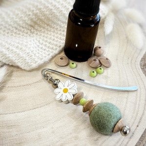 May include: A silver metal bookmark featuring a decorative charm with a white daisy bead, small light green beads, and a larger sage green felt ball. The bookmark is accompanied by a dark amber glass bottle and several loose wooden beads.