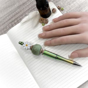 Può includere: Una penna verde e argento con un ciondolo a margherita e una pallina di feltro verde è appoggiata su un quaderno a righe. Sono presenti anche una piccola bottiglia, perline di legno e una piuma. La penna è tenuta da una persona con unghie curate.