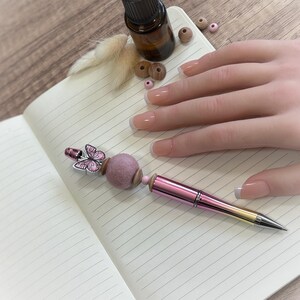 Può includere: Una penna rosa e oro con un ciondolo a farfalla e una pallina di feltro è appoggiata su un quaderno aperto. La penna ha una sfumatura di colore e una punta argentata. Sono visibili anche una piccola bottiglia e perline di legno.
