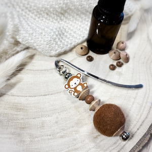 May include: A metal bookmark featuring a monkey charm, wooden beads, and a large brown felt ball. The monkey charm is white and brown with a smiling face. The bookmark is attached to a silver-toned metal hook and includes several small wooden beads.