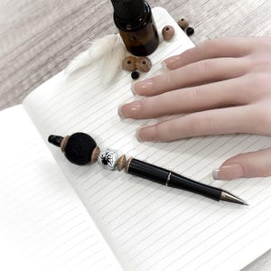 Può includere: Una penna nera con una palla decorativa in feltro e una perlina a forma di ragnatela poggia su un quaderno a righe. Sono presenti anche una piccola bottiglia di olio essenziale e perline di legno. Accessorio di scrittura a tema Halloween.