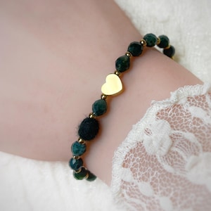 Könnte beinhalten: Ein Armband mit facettierten dunkelgrünen Steinen, kleinen goldfarbenen Metallperlen, einer einzelnen schwarzen, flauschigen Kugelperle und einem glatten, goldfarbenen Herzanhänger in der Mitte. Das Armband ist auf einem flexiblen Band aufgezogen.