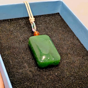 May include: A rectangular, polished green jade pendant with a cream-colored cord. The pendant is suspended from a cord with a small orange bead. The pendant is displayed in a blue box with a black textured interior.