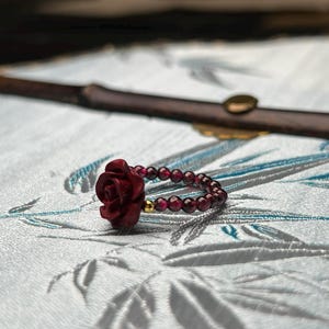 May include: A ring featuring a dark red carved rose focal point and a band composed of small, round, deep red garnet beads. A single small, polished gold-tone metal bead is positioned next to the rose, accenting the circular beaded band.