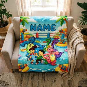Puede incluir: Una manta azul personalizada con una escena de piscina con dragones y niños de dibujos animados. La palabra "NAME" se muestra en letras azules grandes. También se ven palmeras y pistolas de agua en el diseño. La manta está sobre un sillón beige.