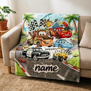 May include: A colorful blanket featuring cartoon cars racing on a track. The design includes a brown tow truck, a red race car, and a blue car, with the word "name" in a speech bubble. Palm trees and checkered flags are also visible.