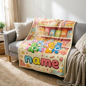 Op de afbeelding: Een kleurrijke deken met een cartoon snoepwinkelscène met Care Bears. De deken heeft een lichtbeige achtergrond met een snoepwinkelontwerp, Care Bears en het woord "name" in grote, kleurrijke letters. De deken ligt over een grijze bank.