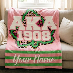 May include: Pink plush blanket featuring the text "AKA 1908" in white, outlined in green, with a leopard print design in pink, green, and yellow. The bottom has green and pink stripes with the text "Your Name" in white.