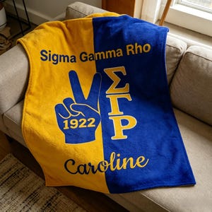 Könnte beinhalten: Eine gelb-blaue Fleece-Decke mit dem Text "Sigma Gamma Rho", einem Peace-Zeichen mit "1922" und dem Namen "Caroline". Die Decke liegt über einem beigen Sofa.